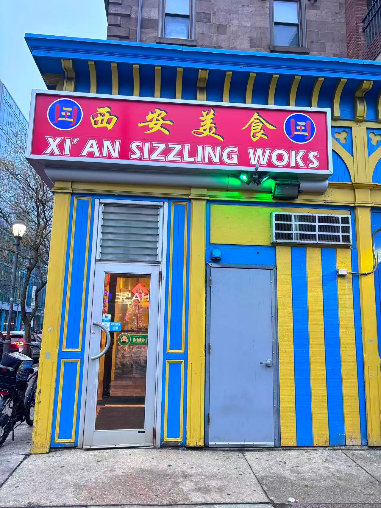The exterior of Xi'an Sizzling Woks, featuring its vibrant red, blue, and yellow storefront with bilingual signage.